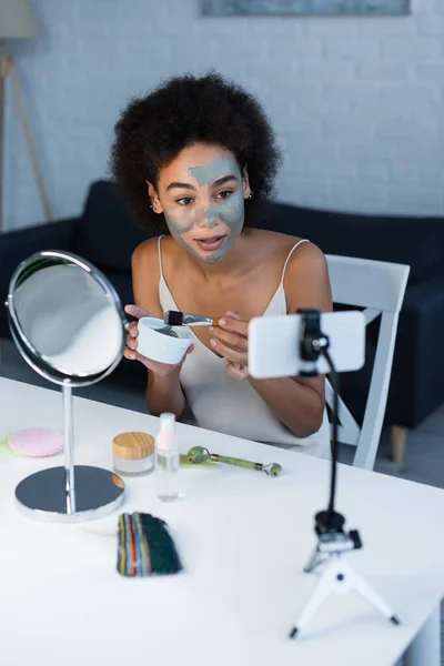 Blogueiro afro-americano segurando máscara de barro e escova cosmética perto do smartphone em casa — Fotografia de Stock