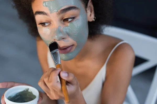 Joven mujer afroamericana aplicando máscara de arcilla en la cara en casa - foto de stock