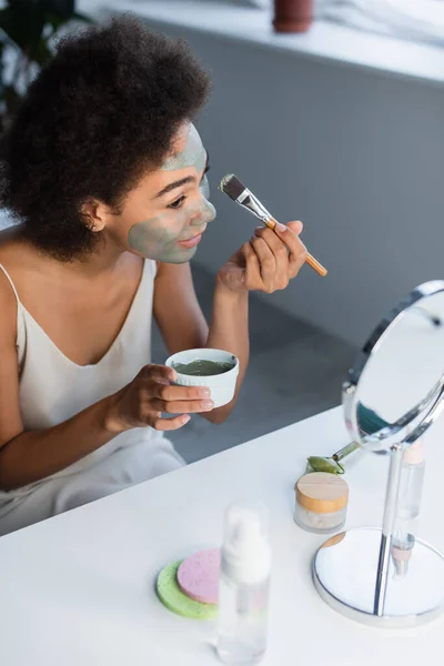 Mujer afroamericana aplicando mascarilla de arcilla cerca de crema cosmética, rodillo de jade y espejo en casa - foto de stock