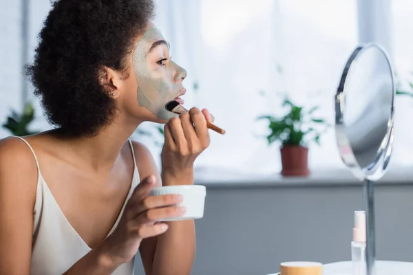 Vista lateral de mujer afroamericana aplicando mascarilla cerca del espejo en casa — Stock Photo