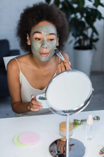 Mujer afroamericana aplicando mascarilla cerca de espejo y rodillo de jade en casa - foto de stock