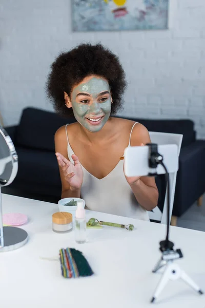 Blogueur afro-américain souriant avec masque facial regardant smartphone à la maison — Photo de stock