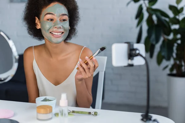 Femme afro-américaine souriante avec masque en argile tenant une brosse cosmétique près du téléphone portable à la maison — Photo de stock