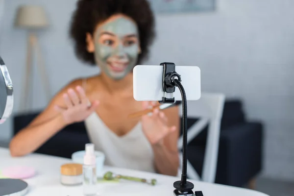 Smartphone cerca borrosa blogger afroamericano con máscara de arcilla en la cara en casa - foto de stock