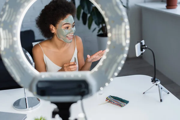 Blogger afroamericano sonriente con máscara de arcilla en la cara sosteniendo cepillo cosmético cerca de teléfono inteligente y anillo de luz - foto de stock