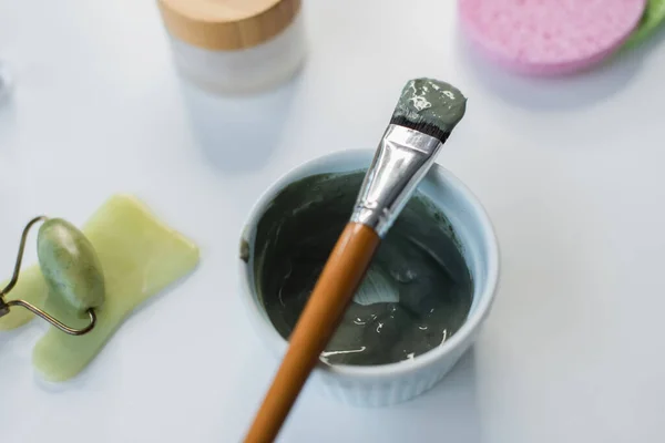 Top view of cosmetic brush on clay mask near jade roller and cosmetic cream on table — Fotografia de Stock