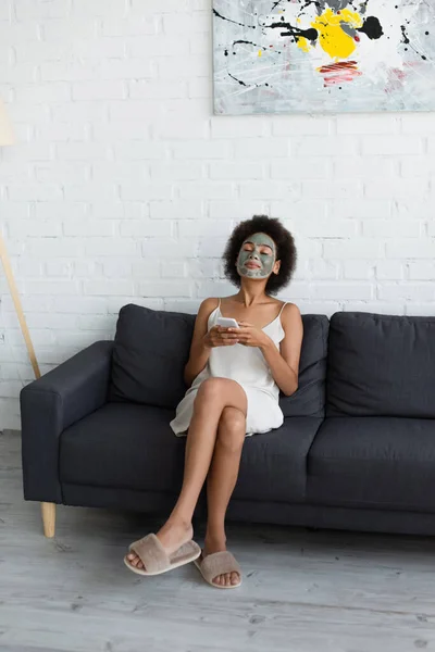 Jeune femme afro-américaine avec masque en argile sur le visage en utilisant un smartphone sur le canapé à la maison — Photo de stock