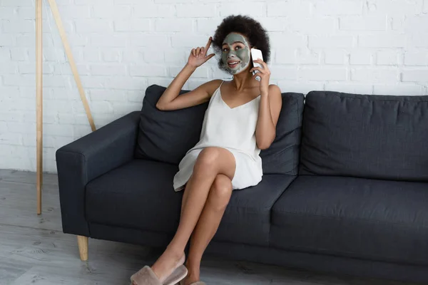 Sorridente donna afroamericana con maschera di argilla sul viso che parla su smartphone sul divano di casa — Foto stock