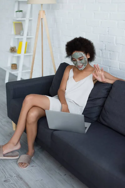 Sourire femme afro-américaine avec masque en argile sur le visage ayant appel vidéo sur ordinateur portable à la maison — Photo de stock