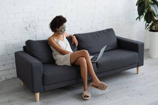 Jeune femme afro-américaine avec masque en argile sur le visage en utilisant un ordinateur portable sur le canapé à la maison — Photo de stock