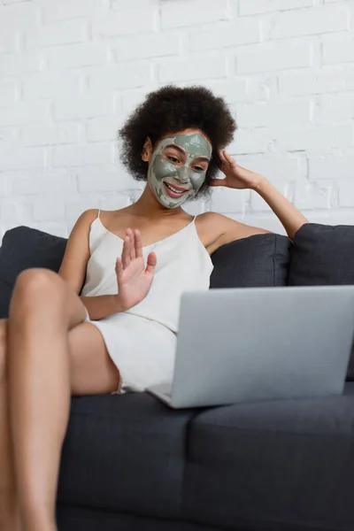Jeune femme afro-américaine avec masque en argile sur le visage ayant chat vidéo sur ordinateur portable à la maison — Photo de stock
