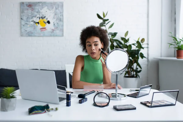 Femme afro-américaine appliquant blush près de cosmétiques décoratifs, miroir et ordinateur portable à la maison — Photo de stock