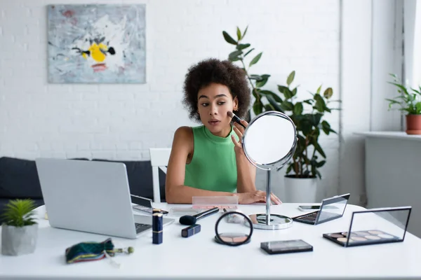 African american woman applying blush near decorative cosmetics, mirror and laptop at home — Foto stock