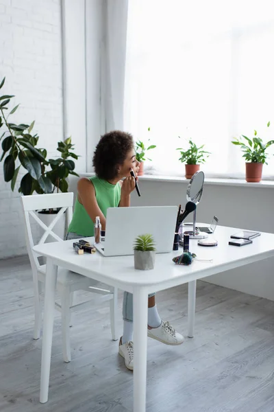 Vista lateral da mulher afro-americana aplicando blush perto de dispositivos e cosméticos decorativos em casa — Fotografia de Stock