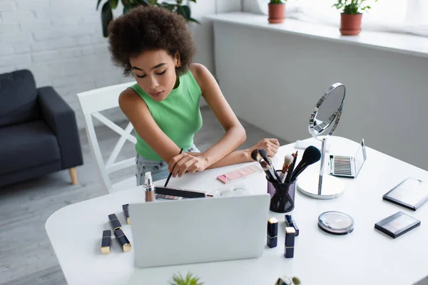 African american woman holding cosmetic brush near eye shadows and gadgets at home — Foto stock