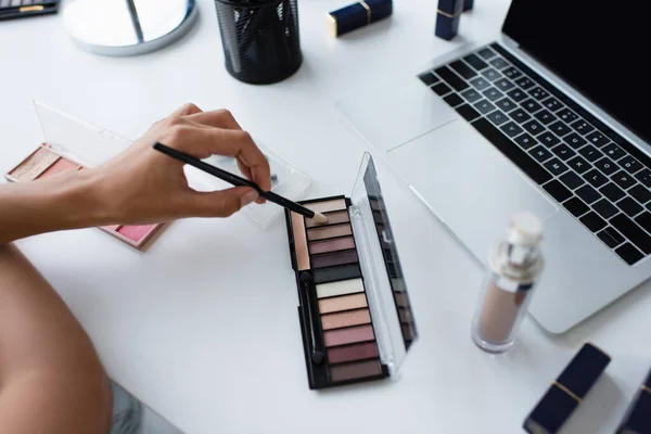 Vue recadrée d'une femme afro-américaine tenant une brosse cosmétique près des ombres à paupières et d'un ordinateur portable à la maison — Photo de stock