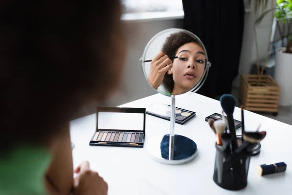 Mujer afroamericana borrosa aplicando sombra de ojos cerca del espejo y pinceles cosméticos en casa - foto de stock