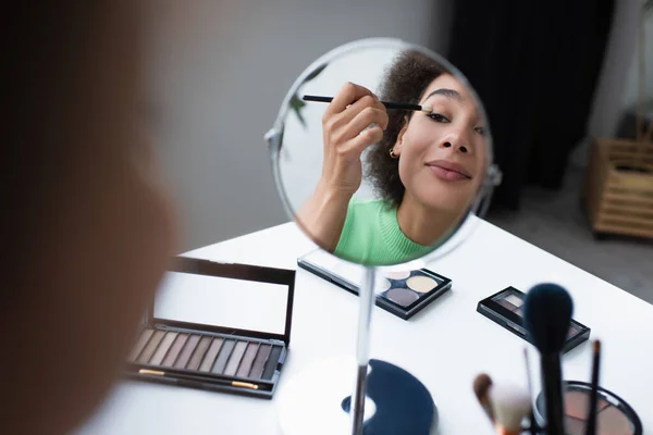 Mujer afroamericana sonriente aplicando sombra de ojos cerca del espejo y pinceles cosméticos borrosos en casa - foto de stock