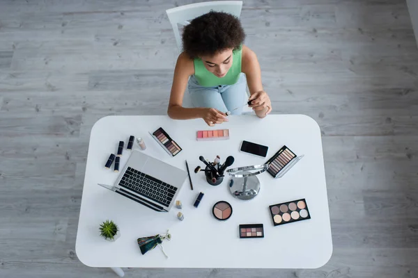 Vista aerea della donna afro-americana che tiene balsamo labbra vicino a dispositivi e cosmetici sul tavolo — Foto stock