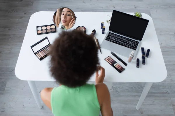 Vue aérienne de la femme afro-américaine appliquant un gel frontal près du miroir et de l'ordinateur portable à la maison — Photo de stock