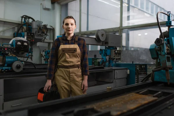 Welder in overalls holding welding mask and looking at camera in factory — Stock Photo