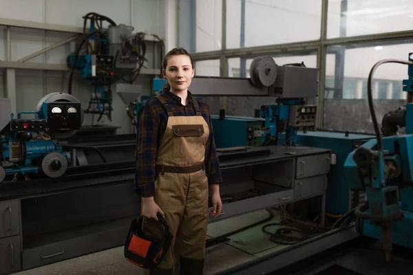 Young welder holding protective mask and looking at camera in factory — Stock Photo