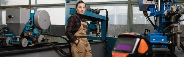 Jeune soudeur regardant la caméra près du masque de soudage et des machines dans l'usine, bannière — Photo de stock