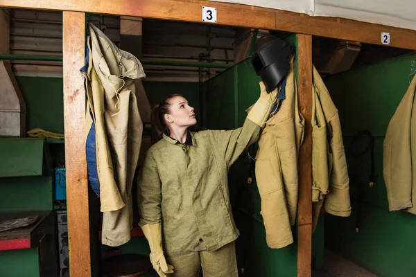 Soldador morena en uniforme tomando máscara protectora en vestidor de fábrica - foto de stock