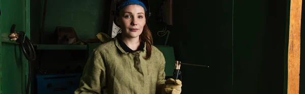 Brünettschweißer schaut in die Kamera, während er Schweißbrenner in der Fabrik hält, Banner — Stockfoto