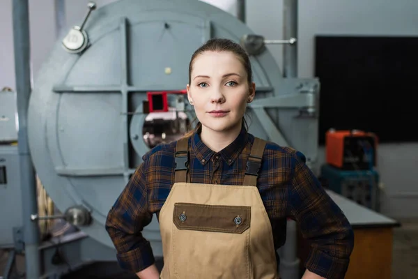 Brunette welder in overalls looking at camera near blurred vacuum laser welding machine in factory — Foto stock