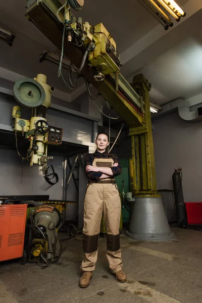 Pleine longueur de soudeur en salopette traversant les bras et regardant la caméra près des machines dans l'usine — Photo de stock