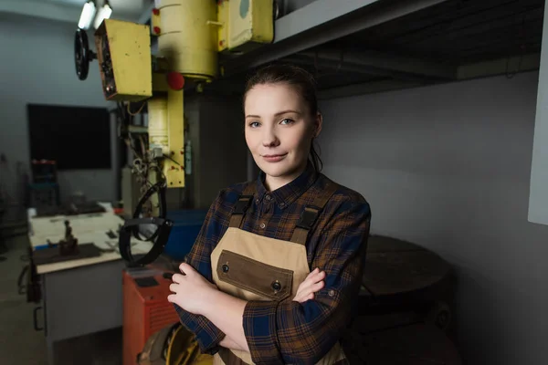 Pretty welder in overalls looking at camera near blurred welding machine in factory — Foto stock