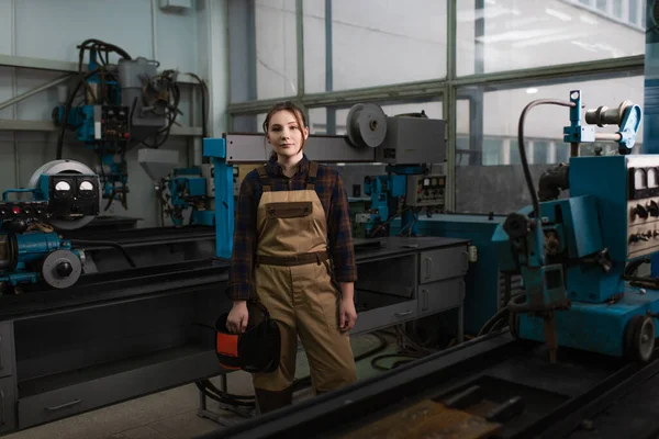 Brunette welder holding protective helmet near welding machines in factory — Foto stock