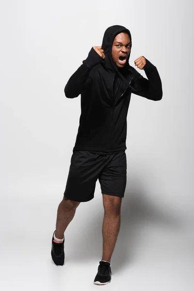 Tutta la lunghezza dell'uomo afroamericano arrabbiato in felpa con cappuccio che combatte sul grigio — Foto stock