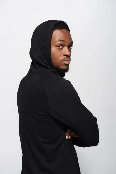 Joven afroamericano en sudadera con capucha mirando a la cámara mientras posa con los brazos cruzados aislados en gris - foto de stock