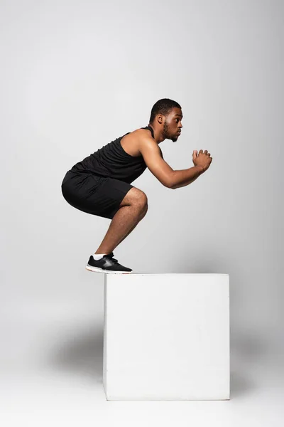 Volle Länge des afrikanisch-amerikanischen Sportlers, der auf White Cube auf Grau springt — Stockfoto