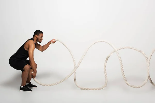 Side view of african american sportsman exercising with battle ropes on grey — стоковое фото