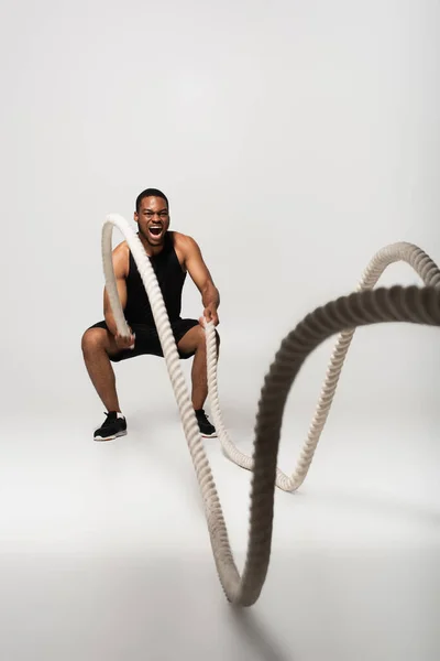 Tutta la lunghezza di sportsman afroamericano urlando durante l'esercizio con corde da battaglia su grigio — Foto stock