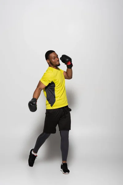 Полная длина сильного африканского спортсмена в боксёрских перчатках, прыгающего на сером — стоковое фото