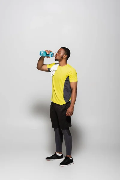 Full length of african american sportsman drinking water from sports bottle on grey — Stock Photo