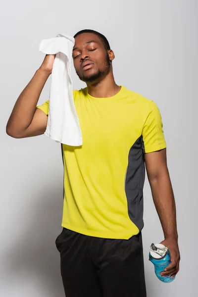 Tired african american sportsman wiping sweat with towel and holding sports bottle on grey — Stock Photo