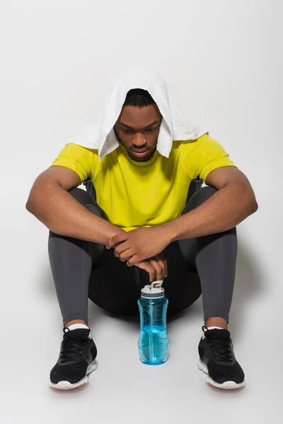 Full length of tired african american sportsman with towel on head sitting near sports bottle on grey — Stock Photo