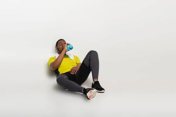 Full length of african american sportsman drinking water from sports bottle while sitting on grey — стоковое фото