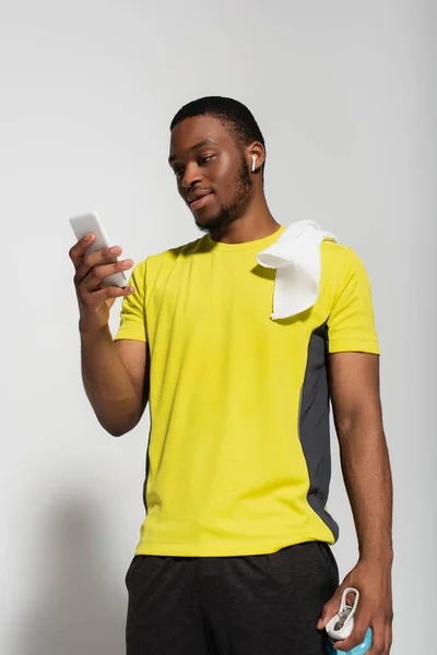Deportista afroamericano con auricular en la oreja usando teléfono inteligente en gris - foto de stock