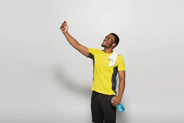 Deportista afroamericano feliz con auricular en la oreja tomando selfie en gris - foto de stock
