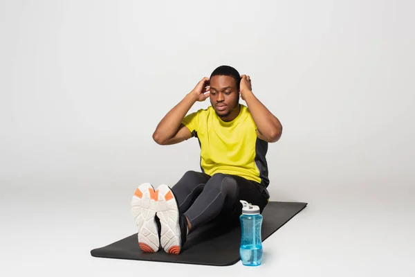 Full length of african american sportsman doing abs exercise on fitness mat near sports bottle on grey — стоковое фото