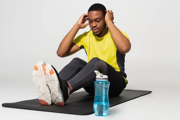 Full length of sportive african american man doing abs exercise on fitness mat near sports bottle on grey — стоковое фото