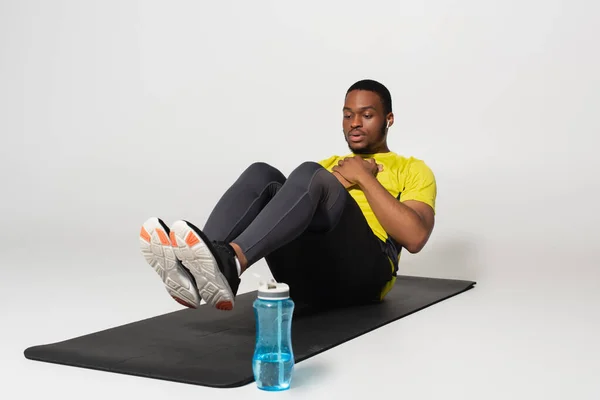 Full length of sportive african american man in earphones doing abs exercise on fitness mat near sports bottle on grey — стоковое фото