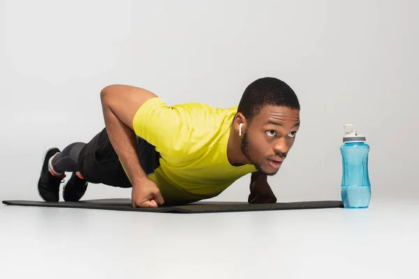 Hombre afroamericano deportivo en auriculares haciendo flexiones en la colchoneta de fitness cerca de la botella de deportes aislado en gris - foto de stock