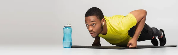 Спортивний афроамериканський чоловік в навушниках робить підштовхування до фітнес-мату біля спортивної пляшки на сірому, банер — стокове фото